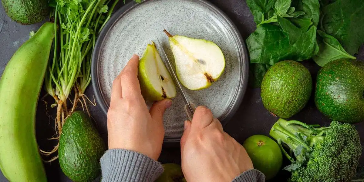 Slicing a pear surrounded by avocados, broccoli, and bananas